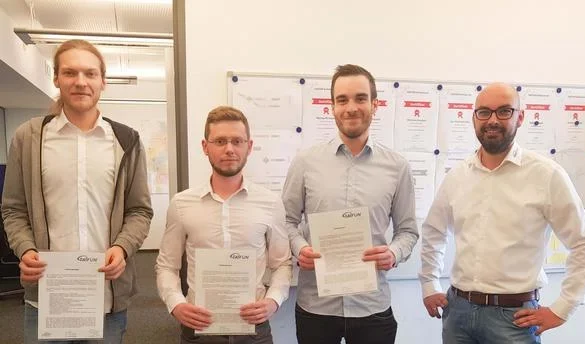 Drei Männer zeigen stolz ihre neuen Zertifikate, ein Kollege steht dabei. Im Hintergrund hängt eine Infotafel mit Unterlagen.