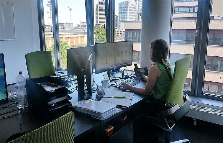 Jemand arbeitet am Schreibtisch, prüft Unterlagen und tippt am Rechner, während draußen die Stadt zu sehen ist.