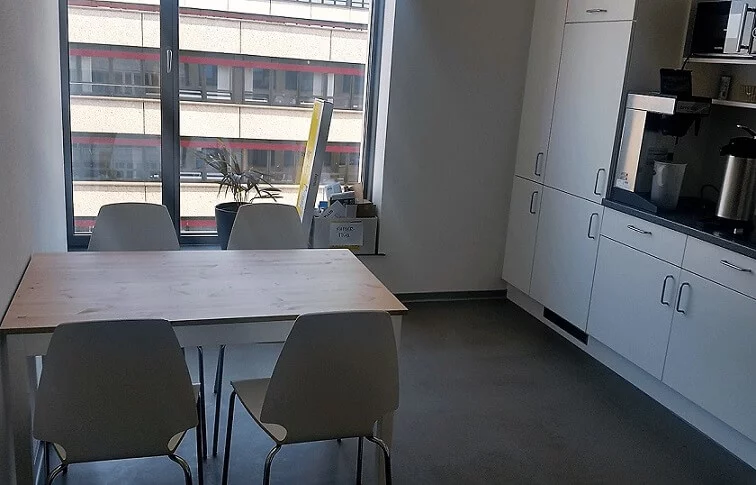 Jemand brüht Kaffee auf, während Kollegen am Tisch über das nächste Projekt sprechen. Helle Küche mit Fenster zum Nachbarhaus.