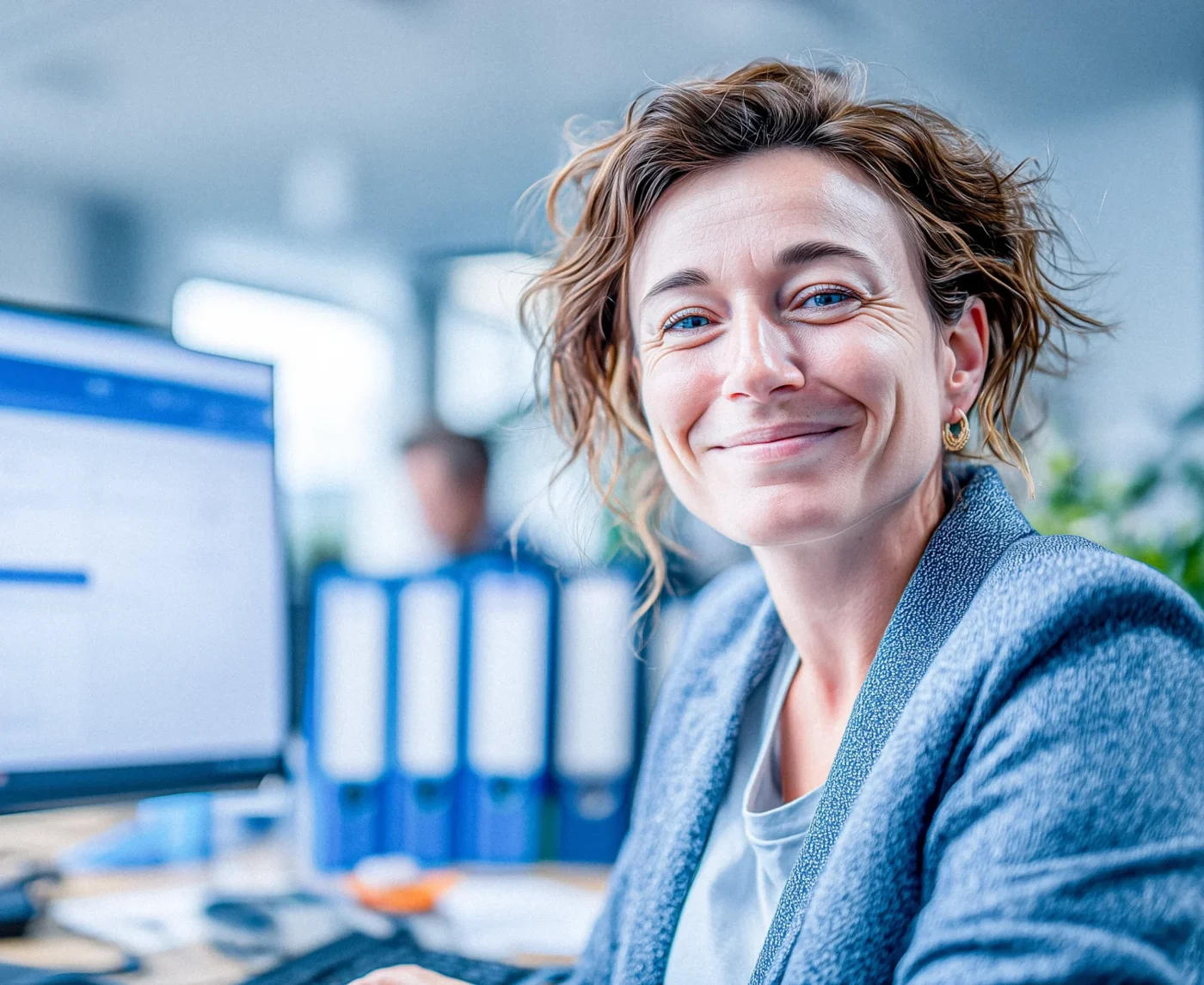 Eine Frau sitzt in einem Büro am Schreibtisch und lächelt in die Kamera.